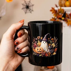 May include: A black ceramic mug featuring a colorful graphic of a white ghost wearing a purple witch hat, a smiling orange jack-o'-lantern, and a small black cat, all surrounded by autumn leaves and flowers in shades of yellow, red, and brown.