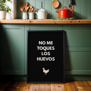 Puede incluir: Un cuadro negro con texto blanco que dice "NO ME TOQUES LOS HUEVOS" y una pequeña ilustración de un pollo. El cuadro se muestra en un entorno de cocina con utensilios de madera y una olla roja.