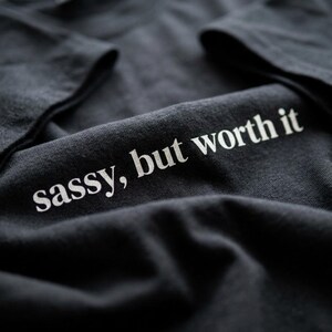 Puede incluir: Camiseta gris oscuro con la frase "sassy, but worth it" impresa en blanco. El texto está centrado en la parte inferior derecha de la camiseta. La tela está ligeramente arrugada y las mangas son visibles en el fondo.