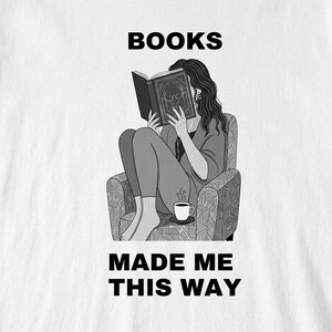 Puede incluir: Camiseta gráfica en blanco y negro con una persona leyendo un libro en un sillón estampado. El texto "BOOKS MADE ME THIS WAY" está impreso encima y debajo de la ilustración. Una taza de café está cerca.