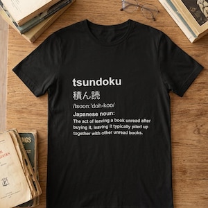 Puede incluir: Una camiseta negra de manga corta con texto impreso en blanco que define el sustantivo japonés tsundoku, incluyendo los caracteres japoneses, la pronunciación fonética y la definición en inglés sobre el acto de dejar libros sin leer.