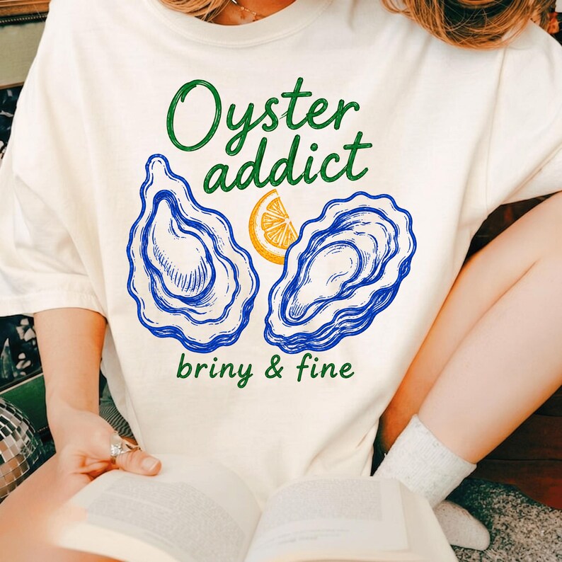 May include: An off-white t-shirt featuring a graphic print of two blue oysters and a yellow lemon slice. The text reads Oyster addict in green above the oysters and briny & fine in green below the oysters. The design is centered on the chest.