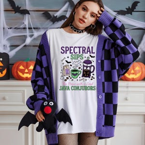 Puede incluir: Una camiseta blanca con un estampado gráfico con el texto "SPECTRAL SIPS" y "JAVA CONJURORS" junto a ilustraciones de una cafetera de émbolo, una taza, una bolsa de café, murciélagos y huesos, bajo un cárdigan de punto a cuadros morados y negros.