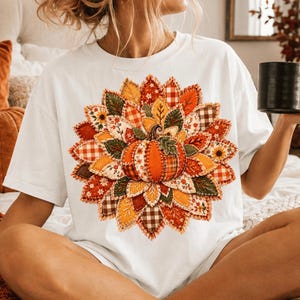 Op de afbeelding: Een wit t-shirt met korte mouwen en een grote, ronde afbeelding van een centrale oranje pompoen, omringd door lagen gestileerde bladeren in diverse herfstkleuren en patronen, waaronder rood, geel, groen, ruitjes en bloemenprints.