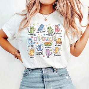 Puede incluir: Una camiseta blanca de manga corta con un gráfico colorido de varios gatos de dibujos animados y el texto "IT'S OKAY TO", "MAKE MISTAKES", "AVOID EYE CONTACT", "NEED ROUTINES", "BE YOURSELF", "FEEL DEEPLY", "TAKE BREAKS", "NEED SPACE", "UNMASK", "COMMUNICATE DIFFERENTLY", "STIM" y "BE DIFFERENT."