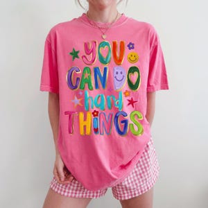 May include: A pink short-sleeve t-shirt featuring the colorful text YOU CAN DO hard THINGS in a bold, multi-colored font. The design includes various graphic elements such as a yellow smiley face, small flowers, and stars in green, red, and blue.