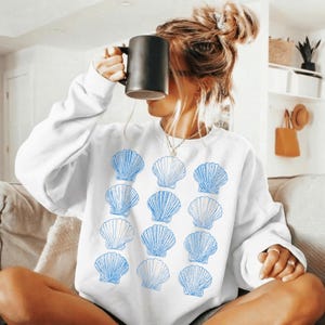 Puede incluir: Sudadera blanca con un patrón de ilustraciones de conchas marinas azules. La sudadera es de cuello redondo y parece estar hecha de un material suave. El tema general es costero o de inspiración playera.