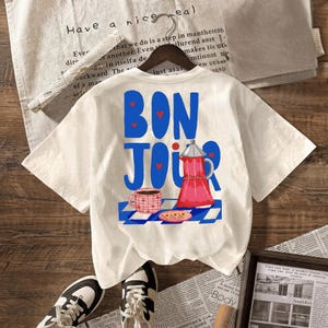 May include: A cream-colored short-sleeve t-shirt featuring a graphic print with the blue text BONJOUR, small red hearts, a red moka pot, a pink patterned coffee mug, and a plate with heart-shaped cookies on a blue and white checkered tablecloth.