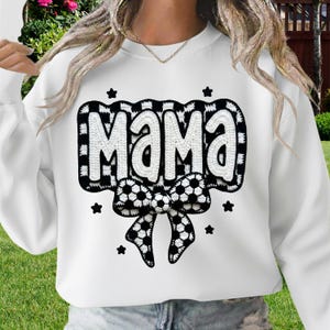 May include: White sweatshirt with the word "MAMA" in a black and white checkered design. A black and white soccer ball bow is below the word, with black star accents. A casual top for mothers.
