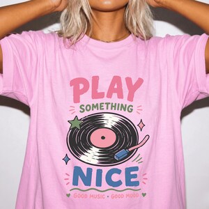 May include: A light pink t-shirt featuring a graphic of a black vinyl record with a pink center. The text reads PLAY SOMETHING NICE and GOOD MUSIC GOOD MOOD. The design includes small green and blue stars, plus pink and green decorative accents.