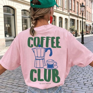 May include: A light pink t-shirt featuring a dark green graphic on the back that reads COFFEE CLUB in bold letters. The design includes a blue line-art illustration of a moka pot and a small coffee cup on a saucer, with text reading EST. IN ITALY / SINCE 1919.