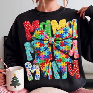 May include: Black sweatshirt featuring the word "MAMA" in various patterned fonts. A large bow made of jigsaw puzzle pieces is the main design. A white mug with a Christmas tree design is also visible.