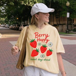 May include: A cream-colored t-shirt featuring a graphic print of four red strawberries and three small white flowers with blue centers. The text "Berry Happy" is printed in red above the fruit, and "sweet days ahead" is in green below.