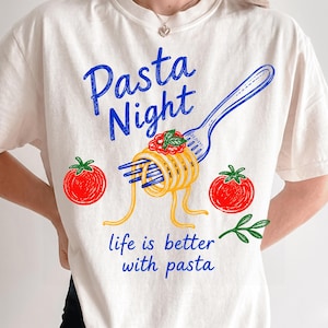 May include: An off-white t-shirt featuring a blue graphic of a fork twirling yellow spaghetti with red sauce and green basil. Two red tomatoes are on either side. The text reads Pasta Night and life is better with pasta in blue script.