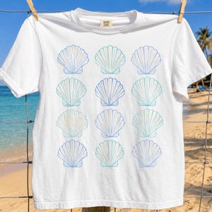 Puede incluir: Una camiseta blanca de manga corta con un patrón de cuadrícula repetitivo de doce ilustraciones de conchas marinas en tonos azul y verde azulado. La prenda tiene cuello redondo y una etiqueta en el cuello interior que dice Comfort Colors.