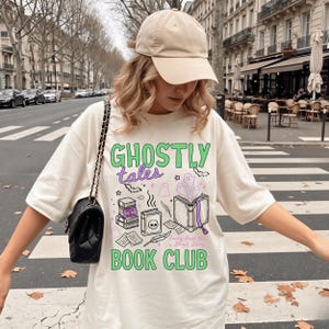 May include: An off-white t-shirt featuring a graphic print with the text "GHOSTLY tales BOOK CLUB" and "Every Book Has a Ghost Story". The design includes illustrations of books, a skull, ghosts, bats, and a quill pen. A beige baseball cap is worn.