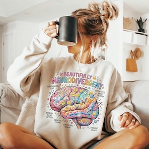 May include: A cream-coloured crewneck sweatshirt featuring a colourful graphic of a brain with the text "BEAUTIFULLY NEURODIVERGENT" and labels including "OVERTHINKING ZONE," "HYPERFOCUS MODE FACTORY," "RANDOM IDEAS FACTORY," "CREATIVE STORM," "EMOTIONAL RADAR," and "THOUGHT CHAOS."