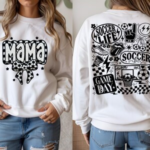 May include: White sweatshirt with a black and white soccer ball design. The front features the word "MAMA" and a bow. The back has soccer-themed graphics and the phrases "SOCCER LIFE", "SOCCER VIBES", and "GAME DAY".