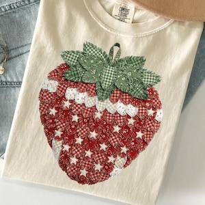 Puede incluir: Camiseta Comfort Colors color crema con un gran diseño de fresa aplicado. La fresa está hecha de tela estampada roja y blanca con detalles de estrellas blancas y hojas verdes. La camiseta tiene cuello redondo.