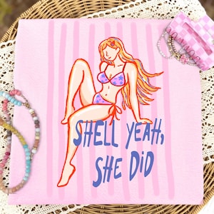 May include: A light pink square napkin featuring vertical stripes and a graphic illustration of a person with long hair wearing a star-patterned bikini. The text "SHELL YEAH, SHE DID" is printed in blue below the figure. Beaded jewelry is visible.