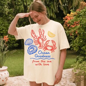 May include: A cream-colored t-shirt featuring a graphic print of a red lobster, a red crab, a blue seashell, a blue oyster, and a yellow lemon slice. The text reads Ocean Goodness in blue and from the sea with love in red below the graphics.
