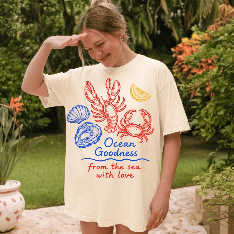 May include: A cream-colored t-shirt featuring a graphic print of a red lobster, a red crab, a blue seashell, a blue oyster, and a yellow lemon slice. The text reads Ocean Goodness in blue and from the sea with love in red below the graphics.