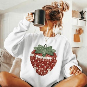 Könnte beinhalten: Weißes Sweatshirt mit einem großen Erdbeer-Design. Die Erdbeere hat einen roten Körper mit weißen Sternen und eine grün-weiß karierte Blattkrone. Eine schwarze Tasse wird vor dem Gesicht gehalten.