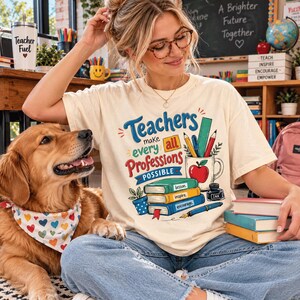 Op de afbeelding: Een crèmekleurig t-shirt met een grafische print met de tekst "Teachers make every all Professions POSSIBLE" naast illustraties van gestapelde boeken met de labels "lesson", "inspire" en "encourage", een mok met een appel en pennen.