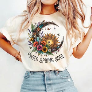 Puede incluir: Camiseta color crema con un diseño floral y solar colorido dentro de una forma de media luna. El diseño incluye flores, abejas y el texto "Wild Spring Soul".