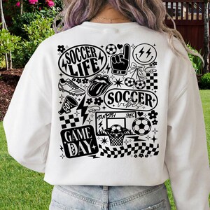 May include: White sweatshirt with a black graphic design featuring soccer-themed illustrations and text. The design includes soccer balls, a hand gesture, lightning bolts, and the phrases "Soccer Life," "Soccer Vibes," and "Game Day."