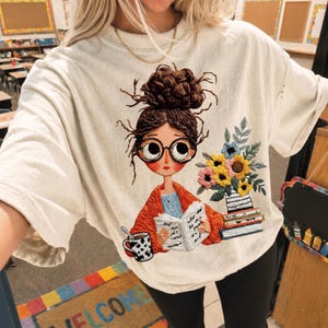 Könnte beinhalten: Ein cremefarbenes T-Shirt mit einer Stickerei, die eine Person mit Dutt und Brille beim Lesen eines Buches zeigt. Das Design umfasst eine Tasse mit Kuhfleckenmuster, einen Bücherstapel und einen bunten Blumenstrauß in einer Vase.
