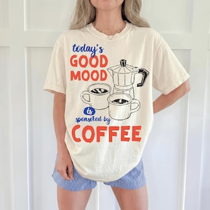 May include: A cream-colored short-sleeve t-shirt featuring a graphic print of a moka pot and two coffee mugs. The text reads today's GOOD MOOD is sponsored by COFFEE in blue and red lettering. The shirt is paired with blue and white striped shorts.