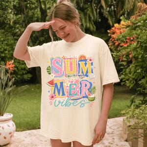 May include: A cream-colored short-sleeve t-shirt featuring a colorful graphic print with the text "SUMMER vibes". The design includes illustrations of a watermelon slice, hibiscus flowers, a surfboard, a coconut drink, and citrus fruit slices.