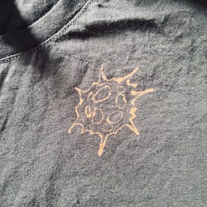 Könnte beinhalten: Dunkelgraues T-Shirt mit Rundhalsausschnitt. Das Shirt hat eine hellbraune, stilisierte Grafik einer Zell- oder Virusform auf der Vorderseite. Das Design hat eine stachelige Umrandung mit mehreren kreisförmigen Formen im Inneren.