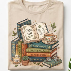 May include: Beige t-shirt featuring a stack of books with titles like "Imagined Realms" and "Nighttime Tales." An open book displays the text "Stories to Keep." A teacup, glasses, a candle, and a succulent are also depicted.