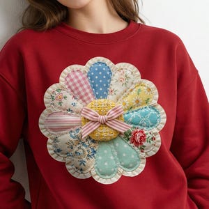 May include: A red sweatshirt featuring a large, appliqued flower design. The flower has various patterned fabric petals in shades of pink, blue, yellow, and green, with a yellow center and a pink striped bow.