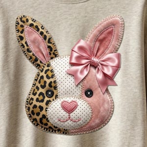 以下が含まれることがあります： ベージュのスウェットシャツには、パッチワークのウサギの顔が描かれています。ウサギはヒョウ柄の耳、ピンクのベルベットの耳、水玉模様の顔をしています。ピンクのサテンのリボンと黒いボタンの目がデザインを完成させます。ハート型の鼻がキュートな印象を添えています。