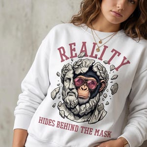 May include: White sweatshirt with a graphic design featuring a monkey wearing pink sunglasses emerging from a broken statue. The word "REALITY" is printed above the graphic, and "HIDES BEHIND THE MASK" is printed below.