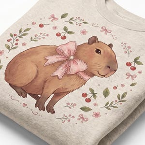 Op de afbeelding: Beige sweatshirt met een illustratie van een capibara met een roze strik. Het ontwerp is omringd door kersen, strikken en bloemmotieven. Het sweatshirt is opgevouwen, wat de zachte stof laat zien.