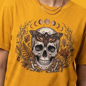 May include: A mustard yellow t-shirt featuring a central graphic of a human skull with a large moth resting on its forehead. The skull is surrounded by a circular arrangement of brown autumn leaves, ferns, mushrooms, and a lunar phase cycle.