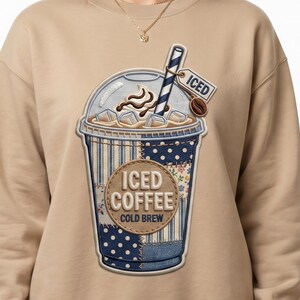 May include: A beige sweatshirt featuring a large, embroidered iced coffee design. The design includes a blue and white striped straw, whipped cream, ice cubes, and a coffee bean. The text "ICED COFFEE COLD BREW" is also visible.