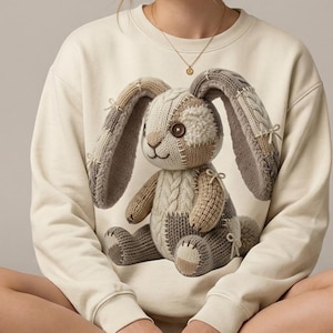 May include: A cream-coloured sweatshirt with a knitted bunny design. The bunny is made from various shades of brown and beige yarn, with long, floppy ears and a button eye. The sweatshirt has ribbed cuffs and a crew neck.