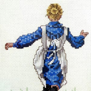May include: Cross-stitch artwork depicting a person in a blue dress and white apron, viewed from the back. The figure has blonde hair and is standing in a field of green grass. The artwork is framed in a light blue border.