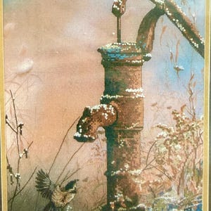 May include: A cross-stitch depiction of a vintage water pump in a winter scene. The pump is brown with white accents, and a small bird perches on top. The background features a soft, muted color palette of blues, pinks, and browns.