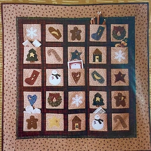 May include: A Christmas-themed quilted wall hanging with 25 pockets, each featuring a different holiday-related applique. The pockets contain small items like a tree, star, snowman, and candy cane. The quilt has a brown and tan color scheme.