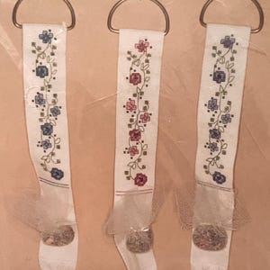 May include: Three white fabric keychains with embroidered floral designs in red, blue, and green. Each keychain has a metal ring at the top and a decorative ball at the bottom. The text "KAPPIE ORIGINALS LTD." is visible at the top.