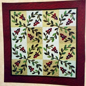 May include: A square quilt featuring a repeating pattern of dark red berries and green leaves on a grid of alternating cream and tan blocks. The entire piece is framed by a wide, solid dark red border with intricate, swirling quilted stitching.