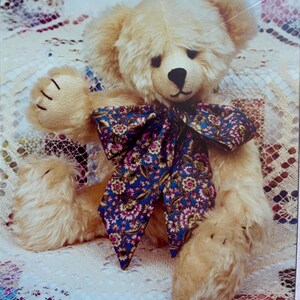 May include: A plush, light brown teddy bear with a dark blue bow featuring a floral pattern in pink and white. The bear has a black nose and stitched claws on its paws. The bear is lying on a patterned surface.