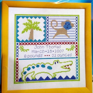 May include: Cross-stitch artwork in a yellow frame. The design features a palm tree, a monkey, and an alligator. Text includes a name, date, and weight: 8 pounds 12 ounces. The artwork uses blue, green, brown, and red colors.