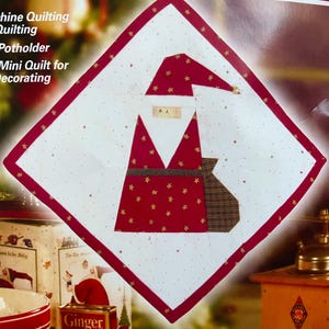 May include: A square Christmas-themed potholder or mini quilt featuring a Santa Claus design. The Santa is depicted in red fabric with gold stars, a white beard, and a brown sack. The potholder has a white background with small gold stars and a red border.